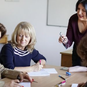 The teacher talking to the adult students sitting around the desk in the classroom.